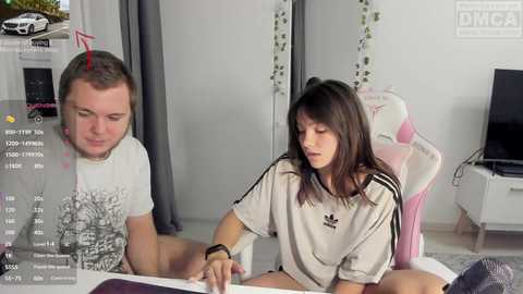 A video of a young woman with long brown hair in a white Adidas shirt, sitting next to a young man with short brown hair in a gray t-shirt, both working on a laptop in a modern, minimalist bedroom.