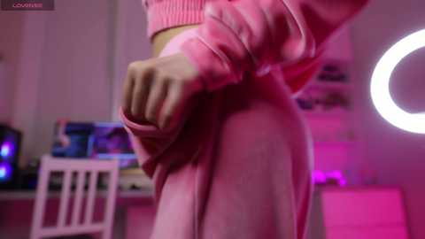 A close-up video of a person wearing a pink sweater, with their hands pulling the sweater up, in a dimly lit room with pink lighting and a ring light.