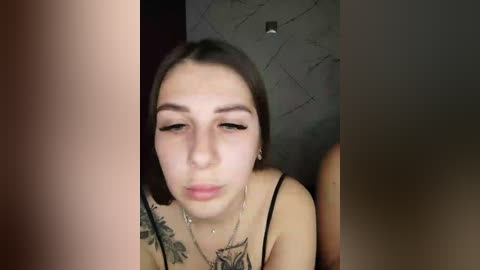 Video of a young Caucasian woman with fair skin, brown hair, and a tattoo on her shoulder, wearing a black tank top. She appears solemn and slightly tilted head. Background includes a dark wall and a faint, blurry image of another person.