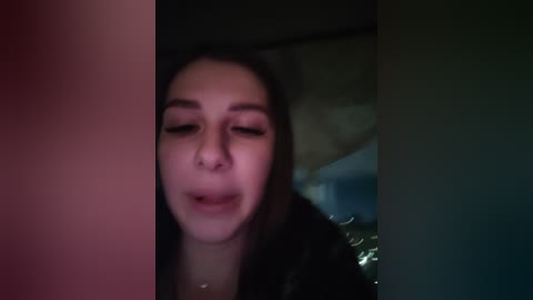 A dimly lit video of a woman with long brown hair, closed eyes, and a serene expression, leaning against a dark, possibly wooden, background.