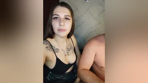 Video of a young woman with long brown hair, fair skin, and medium-sized breasts, wearing a black bra, with visible tattoos on her chest and shoulder. She is seated next to a shirtless man, with a blurred background.