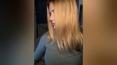 Video of a young Caucasian woman with long, straight, blonde hair, wearing a light gray long-sleeve top, seen from behind in a dimly lit room with dark wallpaper.