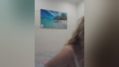 A video of a woman with long blonde hair in a white tank top, standing in a small, dimly lit room with a colorful beach scene poster on the wall.