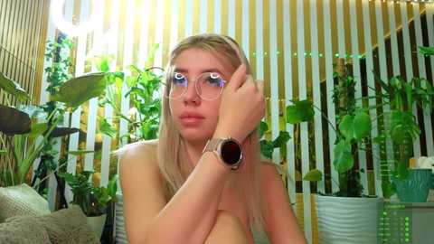 Video of a topless young blonde woman with fair skin, wearing large round glasses and a smartwatch, sitting in a vibrant indoor jungle room with green plants, potted plants, and a vertical striped curtain.