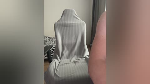 A video of a person sitting on a bed, covered by a gray, striped blanket. The room is dimly lit, with a zebra-print blanket visible on the bed. The image is blurry.