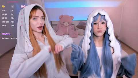 Video of two women in matching light blue hooded robes, one with long, straight orange hair, the other with blue hair, in a cozy bedroom with a teddy bear and soft lighting.