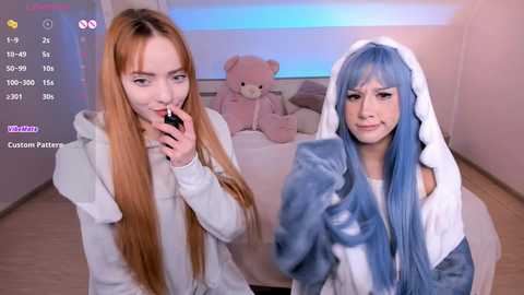 Video of two young women in white hoodies, one with long red hair and the other with long blue hair. Both are holding cell phones. Background features a plush teddy bear and light blue walls.