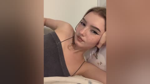 Video of a young woman with fair skin, freckles, and light brown hair, wearing a grey spaghetti-strap top, lying on a bed, partially obscured by a person's legs.