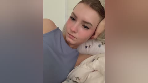 Video of a young woman with light skin and light brown hair, wearing a sleeveless blue top, lying on a bed with a white floral-patterned blanket. Her expression is thoughtful, and the background is out of focus.