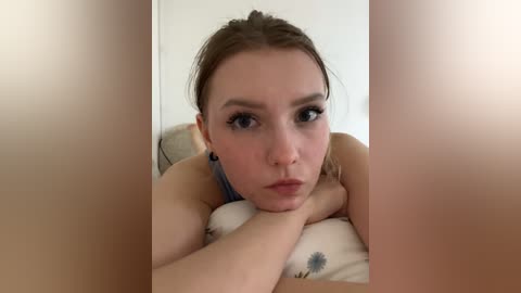 A video of a young Caucasian woman with fair skin, light brown hair tied back, wearing a blue top, lying on a beige pillow, making a serious face.