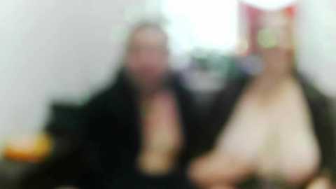 Blurred video of two women with long hair, one with cat ears, both wearing black outfits, sitting indoors with indistinct background.
