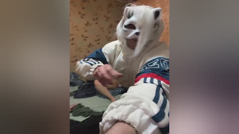 Video of a person in a white bear costume with a large snout, blue and white Adidas tracksuit, and a red arm band, posing in a room with floral wallpaper.