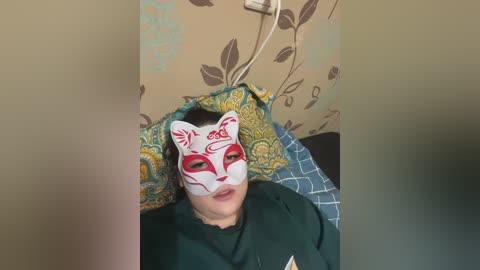 Video of a person wearing a white fox mask with red markings, lying on a bed with a blue quilt. The background features a patterned wallpaper with brown and green leaves.