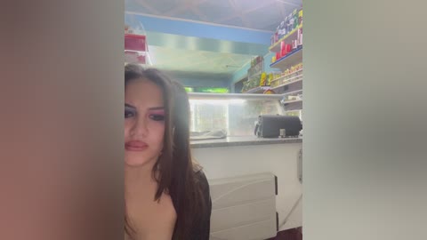 Video of a young woman with long brown hair and light skin, wearing pink eyeshadow, standing in a brightly lit kitchen with shelves of colorful spices in the background.