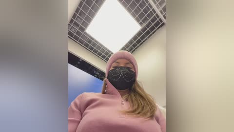 Video of a woman in a pink hoodie and black face mask with rhinestone detailing, standing in a brightly lit office with a grid-patterned ceiling and blue sky visible through a window.