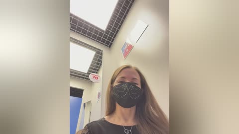 A video of a woman with long brown hair, wearing a black face mask and a black shirt, standing in a dimly lit, tiled hallway with fluorescent lights.