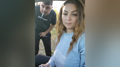 A video showing a young woman with long, wavy brown hair, wearing a light blue sweater, sitting in a car. Behind her, a young man in a black shirt and pants is leaning in.