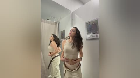 Video of a woman with long dark hair, wearing beige top and pants, standing in a white-walled room, partially reflected in a mirror, with a framed picture on the wall.