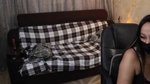 Video of a woman in a black dress, sitting in a black leather chair, with a plaid blanket on a dark wooden couch in a dimly lit room with patterned curtains.