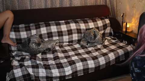 Video of a cozy, dimly lit room with a plaid-covered sofa, a person's bare feet visible on the left, a candle on a side table, and a soft, patterned throw blanket.