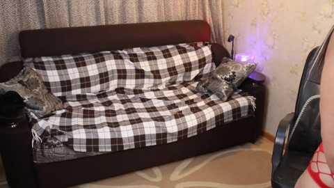 Video of a cluttered bedroom with a plaid-covered bed, patterned pillows, and a chair. A red jacket is partially visible on the floor.