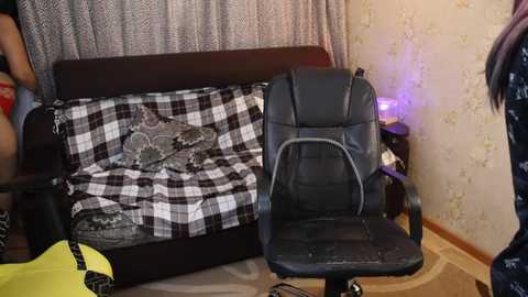 Video of a cluttered bedroom with a black leather office chair, a black-and-white checkered blanket on a wooden bed, and a person in a floral-patterned dress in the background.