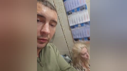 A video shows a young man with short brown hair, wearing a green shirt, looking into a mirror. Behind him, a shelf is filled with white and blue boxes. The image is slightly blurred.