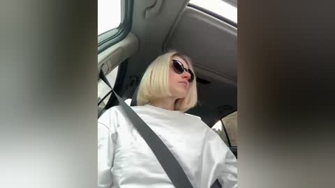 Video of a blonde woman with shoulder-length hair, wearing sunglasses, a white top, and a seatbelt, sitting in a car with a beige interior and sunroof.