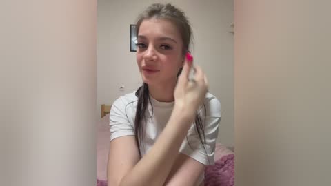 Video of a young woman with light skin, brown hair in braids, wearing a white t-shirt, adjusting her pink earbud with a playful expression.