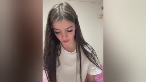 Video of a young woman with long, straight, dark brown hair, wearing a white T-shirt, looking down with a slight smile, standing in a simple, beige-walled room with a small potted plant in the background.