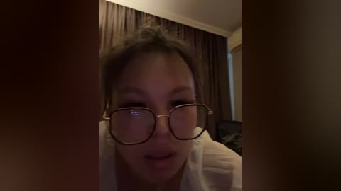 Video of an Asian woman with light skin, dark hair, wearing glasses, a white shirt, and a neutral expression. Background includes dark curtains and a dimly lit room.