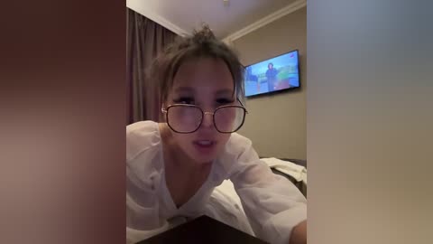 Video of an Asian woman with glasses, light skin, and dark hair, leaning on a bed in a dimly lit room with a TV showing a sports game.