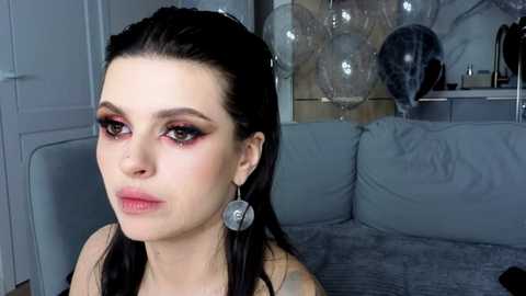 Video of a pale-skinned woman with dark hair, wearing bold pink eye makeup, sitting on a grey couch. Background includes a silver balloon and a decorative pillow.