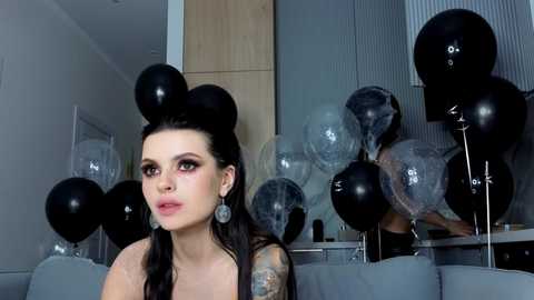 Video of a young woman with dark hair, light skin, and tattoos, wearing black balloons on her head, in a modern, dimly-lit room with gray walls.