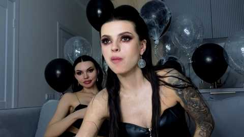 Video of a young woman with long, straight black hair, wearing a black leather corset and large, silver earrings, surrounded by black balloons in a dimly lit room with reflective surfaces.