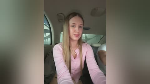 Video of a young woman with long blonde hair, fair skin, and a slender physique, wearing a pink blouse, driving a car with a gray interior.