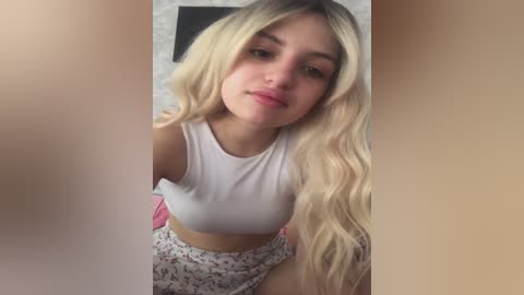 Video of a young Caucasian woman with long, wavy blonde hair, wearing a white crop top and floral skirt, posing indoors with a neutral expression.
