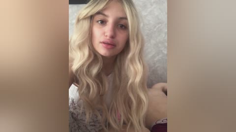 Video of a young Caucasian woman with long, wavy blonde hair and fair skin, wearing a white top, sitting indoors against a beige, textured wall. She gazes directly at the camera with a neutral expression.