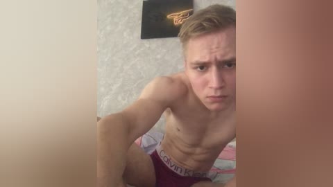 A video of a shirtless, fit young man with fair skin and short blonde hair, wearing maroon Calvin Klein briefs, sitting on a bed with patterned sheets, partially obscured by a beige wall.