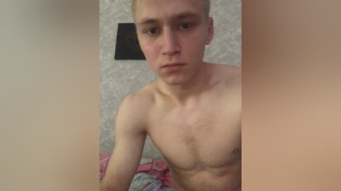 Video of a young, shirtless, pale-skinned man with short blonde hair, looking serious, standing against a floral-patterned wallpaper background.