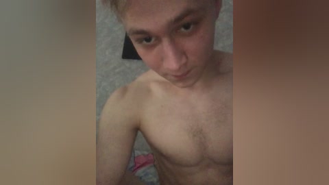 Video of a young, shirtless Caucasian male with pale skin, light brown hair, and a slight beard. He gazes down with a neutral expression. Background includes a light gray carpet and a pink item in the bottom right corner.