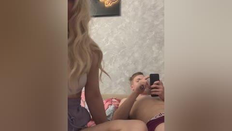 Video of a blonde woman with long hair, wearing a white top, taking a selfie in a bedroom. Background features a man lying on a bed, partially visible, in red underwear.
