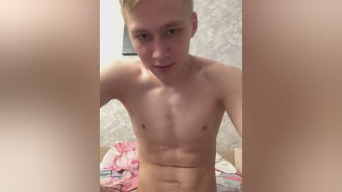 A video of a young, shirtless, fit, white male with short blond hair, standing in a bedroom with a floral-patterned wall and a pink and white bedspread.