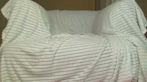 A video of a plush, white, striped armchair covered with a white, knitted blanket, creating a cozy, textured appearance. The background is neutral-toned, with soft lighting highlighting the chair's comfort and warmth.