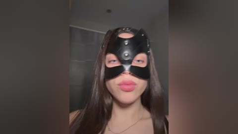 A video of a woman with long, dark hair, wearing a black leather mask with eye holes, pink lipstick, and a gold necklace, standing indoors with blurred background.