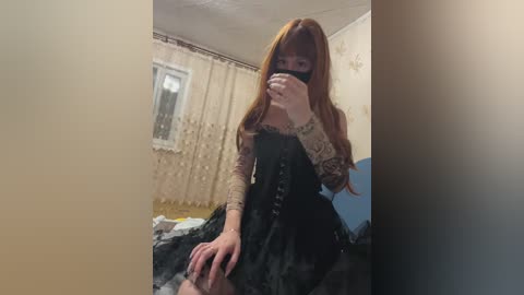 A video shows a young woman with long, auburn hair and pale skin, wearing a black lace dress with intricate detailing, sitting on a bed in a dimly lit, cluttered room.