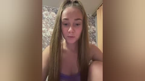 Video of a young, fair-skinned woman with long, wet, blonde hair pulled back, wearing a purple strapless top. Background features a floral-patterned wallpaper and a wooden door.