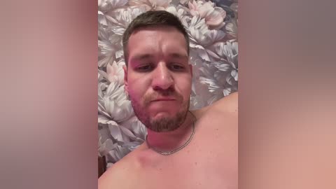 Video of a shirtless man with short brown hair and a beard, lying on a bed of white and pink fake flowers, wearing a silver chain necklace.