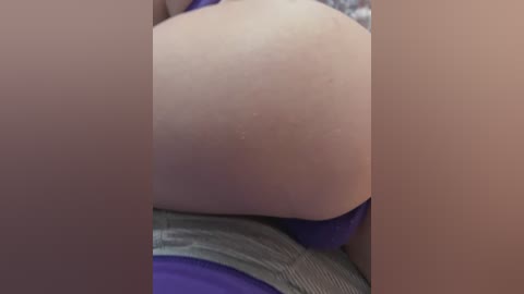 A close-up video shows the bare, slightly bumpy buttocks of a person lying on a purple bedspread, with a gray garment visible at the bottom edge. The skin tone is light.