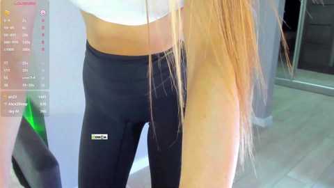Video of a young woman with long blonde hair, wearing tight black leggings and a white crop top, standing in a modern indoor setting.